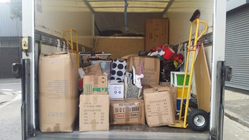 Inside a delivery van parked on a city street, the cargo area is filled with a variety of cardboard boxes and large paper bags, some of which are sealed while others are open or partially collapsed. The boxes are in different sizes, with some bearing printed labels and symbols, indicating reusability or disposal instructions. The boxes and bags appear to be primarily brown or tan, made of corrugated cardboard and paper, with visible creases, tape, and minor dents from handling. On the left side, a large cardboard box rests against the wall of the van, with smaller boxes and bags stacked in front and around it, some labeled with handwritten notes. The right side contains a green plastic container partially visible behind the boxes, and two yellow hand trucks or dollies leaning against the side wall, one with a black cloth draped over it. The environment outside the van suggests an urban setting, with a grey concrete pavement and a metal roller shutter nearby. The scene appears to be related to waste collection or rubbish removal services, such as those offered by Business Waste Removal Peckham, situated in South London, with the image capturing a typical load ready for disposal or recycling.