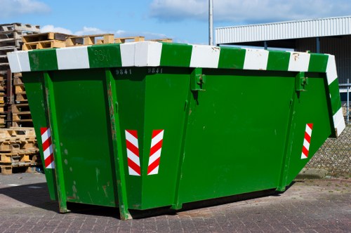A large, rectangular green outdoor rubbish skip with a reinforced metal construction, positioned on a paved surface in an industrial or commercial area. The skip features a slightly sloped top with white and green reflective markings along the edges for visibility. On the side near the top, there is a small black handle or latch, and the skip's vibrant green paint appears clean and well-maintained. In the background, there's a stack of wooden pallets to the left, indicating storage or disposal activity related to waste management, and part of a warehouse or industrial building with a corrugated metal exterior visible behind and to the right. The overall scene suggests a waste collection or disposal operation, typical of services provided by Business Waste Removal Peckham, with neutral natural lighting and no visible waste inside the skip itself, emphasizing its readiness for rubbish collection.
