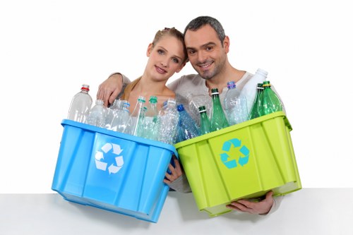 A young couple standing close together, both smiling and looking at the camera, each holding a plastic recycling bin; the woman on the left holds a blue bin and the man on the right holds a green bin, both filled with assorted empty plastic bottles and containers of various sizes, colors, and textures. The plastic bottles include clear, green, and brown colors, with some having caps attached, and they appear to be made from smooth, semi-glossy plastic material. The bins, made of durable plastic with a recycling symbol on their sides, are positioned in front of a plain white background, emphasizing their vibrant colours and contents. The scene suggests a focus on recycling and waste separation, consistent with services provided by Business Waste Removal Peckham in the London Borough of Southwark, especially within the context of environmentally conscious rubbish removal for businesses. The lighting is even, highlighting the clarity and transparency of the plastic bottles, while the overall composition emphasizes responsible waste management practices related to plastic recycling.