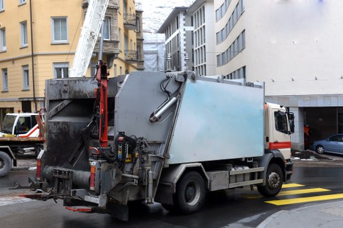 A large commercial waste collection truck parked on a city street, positioned near the curb in front of multi-story buildings with mixed brick and modern facades. The truck's body is primarily light grey with a blueish tint, featuring a metal finish that appears weathered with some visible dirt and grime. On the rear, there is a hydraulic lifting mechanism and equipment for emptying waste containers, and the truck is fitted with a side door. The vehicle occupies a designated parking space marked by yellow and black striped lines on the tarmac, with a driveway or loading area on the right side. In the background, other vehicles including a small van and a car are visible, along with multi-storey residential or commercial structures common in Peckham, London, area. The scene suggests routine rubbish removal operations by Business Waste Removal Peckham, with overcast lighting contributing to a neutral, professional atmosphere suited for urban waste collection services.