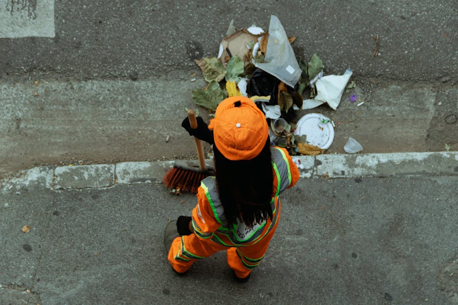 An individual dressed in high-visibility orange workwear with reflective stripes, wearing a matching orange cap, is viewed from above while cleaning a concrete sidewalk. The person is holding a broom 