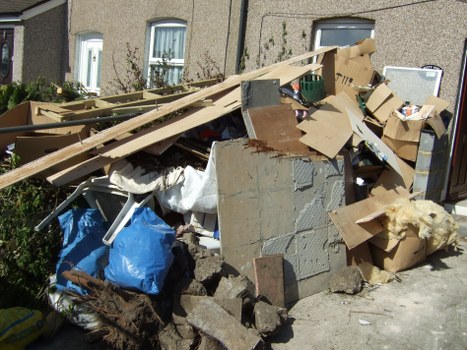 A large pile of mixed waste materials is situated on a paved surface in an outdoor area, likely adjacent to a residential building. The debris includes broken pieces of concrete, bricks, and pavers at the base, with various cardboard boxes, some flattened and others still intact, scattered throughout the pile. There are also several wooden planks and panels, some leaning against the pile or resting on top, exhibiting natural wood textures and light brown hues. A white fabric or cloth is partly draped over section of the waste, and a blue plastic bag is visible among the rubbish. In the background, the side of a residential building with small windows and a garden area with shrubs can be faintly seen, suggesting this may be a domestic or small commercial property in Peckham. The scene is captured under natural daylight, highlighting the textures and disarray of the waste materials. Business Waste Removal Peckham handles such rubbish clearance, including cardboard, construction debris, and household waste.